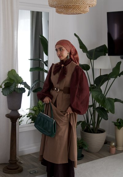 Mujer con atuendo marrón y burdeos y pañuelo en la cabeza sostiene un bolso verde, de pie junto a grandes plantas en macetas y un espejo de pie en una habitación luminosa.