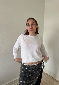 Jeune femme aux cheveux bruns courts portant une chemise blanche à manches longues et une jupe en dentelle noire, debout à l'intérieur contre un mur blanc texturé uni.