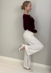 Cardigan bordeaux à manches longues, pantalon blanc à jambes larges et chaussures blanches à talons hauts. Le modèle pose de profil, mettant en valeur les détails des chaussures et du pantalon.