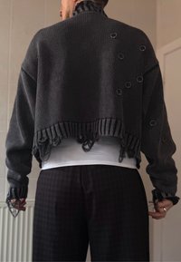 Pull gris court avec un tricot texturé, ourlet effiloché et accents métalliques circulaires. Porté sur un haut blanc et assorti à un pantalon à carreaux foncés.