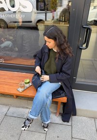 Una donna è seduta su una panchina di legno, indossa un cappotto blu navy sopra un maglione verde e jeans blu, tiene in mano una borsa nera con texture mentre due bevande sono su un vassoio.