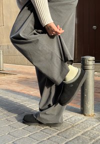 Personne portant un pantalon gris et des chaussures UGG noires à enfiler lève une jambe, tenant le pantalon relevé pour révéler des chaussettes de couleur claire en extérieur.