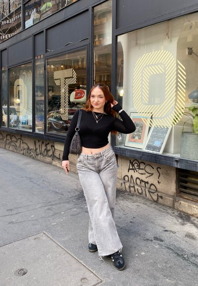 Joven mujer con un top corto negro y unos pantalones de tiro alto grises posando en la acera de la ciudad junto a ventanas con marco negro y grandes letras a rayas O, T, Q.