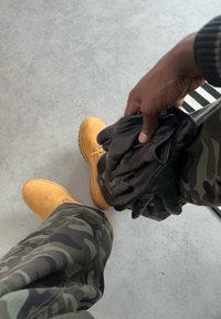 Personne portant un pantalon camouflage et des bottes jaunes tenant des gants en cuir noir sur un sol gris avec le bord d'un tapis rayé.
