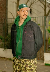Veste en denim foncé avec surpiqûres orange, sweat à capuche vert en dessous, pantalon camouflage, et casquette NY verte. Textures douces et aspect superposé.