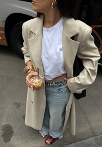 Beżowy oversize'owy płaszcz, biały krótki t-shirt, jasnoniebieskie szerokie dżinsy, pasek w wężowy wzór, trzymając szklankę lodowatej kawy, włożone bordowe sandały.