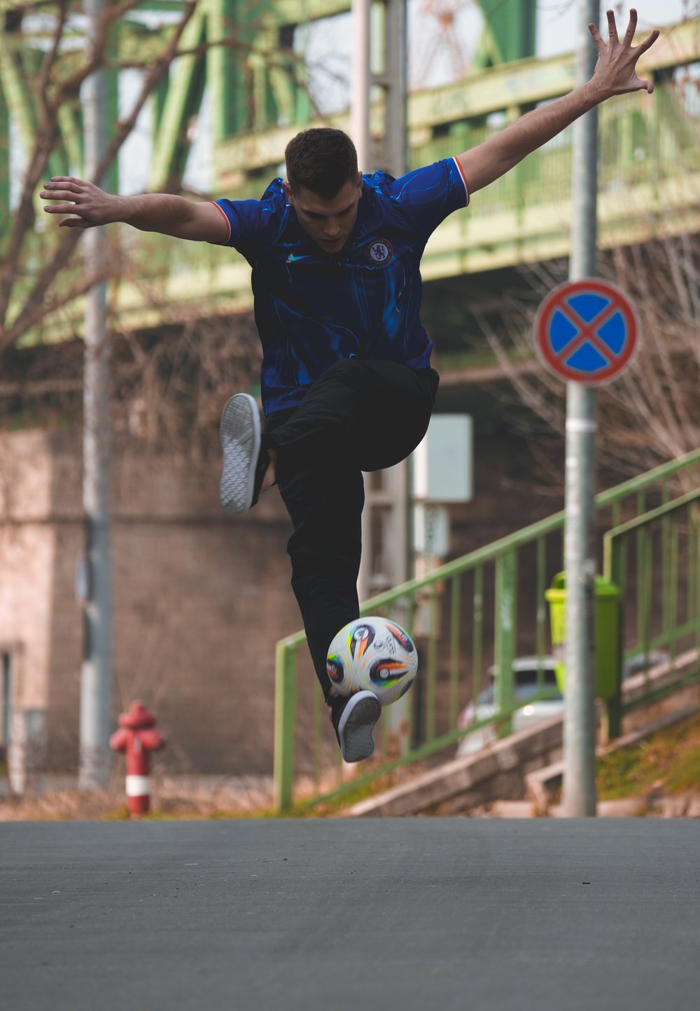 A person wearing a blue graphic football jersey and black trousers is juggling a white football with colourful panels on a street.