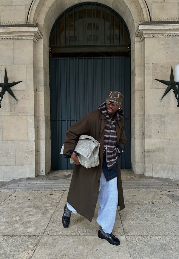 Mann trägt einen langen braunen Mantel, gestreiften Kapuzenpullover, graue Jogginghose und eine Kappe, trägt eine große strukturierte Tasche und geht an einer gewölbten Tür vorbei.