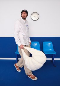 A white fluffy blanket is held by an individual in a white shirt featuring graphic designs and beige trousers, standing in front of blue plastic chairs.