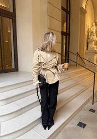 Femme aux cheveux blonds portant une veste beige et un pantalon noir, debout sur des marches en pierre blanche, tenant une tasse de café et un sac noir à l'intérieur d'un bâtiment historique.