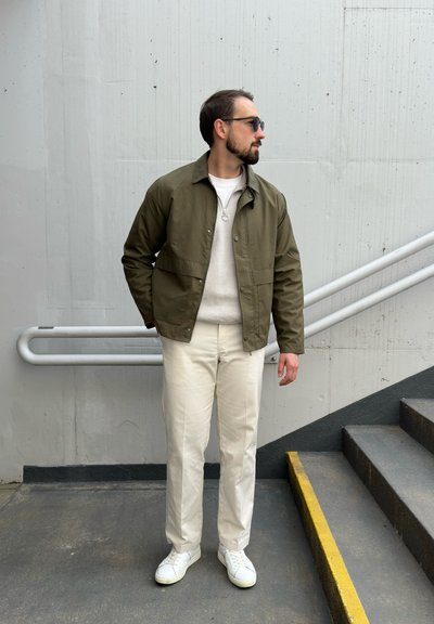 Chaqueta ligera verde olivo sobre un suéter beige, combinada con pantalones color crema y zapatillas blancas, de pie en un vestíbulo.