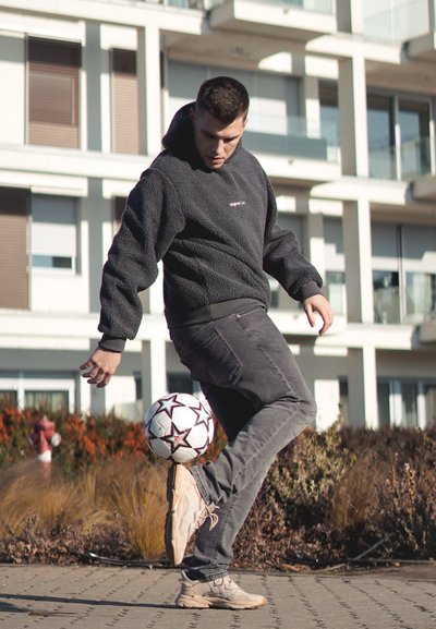 Sudadera con capucha negra, jeans grises y zapatillas beiges; realizando un truco con un balón de fútbol blanco con estrellas negras.