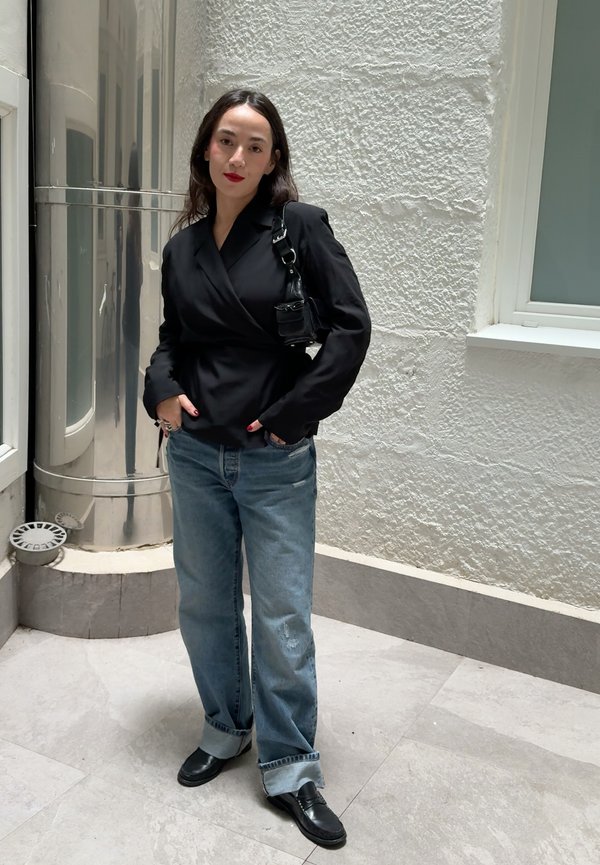 Une femme portant un haut noir portefeuille, un jean bleu à revers et des chaussures noires pose, les mains dans les poches, près d’un mur blanc texturé.