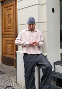 Lichtroze geruite blouse met knopen en een ontspannen pasvorm, gecombineerd met donkere, loszittende broek; grijze beanie; staand tegen een gestructureerde muur.