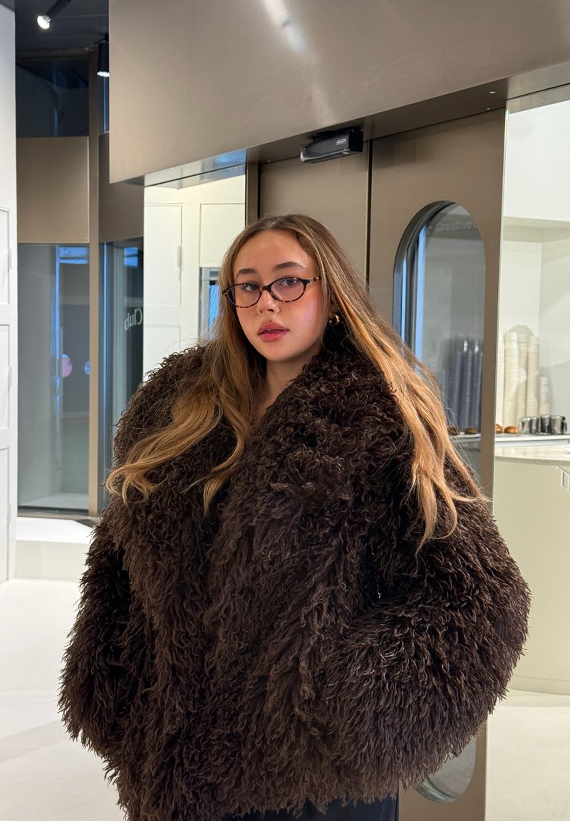 Ung kvinne med langt hår som bærer briller og en stor mørk brun, lurvete frakk innendørs nær en moderne glassdør.