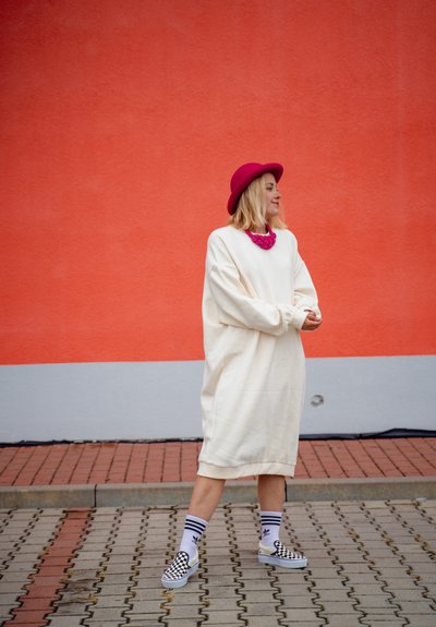 Vestido sudadera blanco oversize con mangas largas, acompañado de un sombrero rosa brillante y un collar chunky rosa. Zapatos slip-on de cuadro blanco y negro.