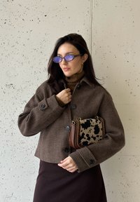 Καφέ καρό cropped μπουφάν με μεγάλους κουμπιά, φορεμένο πάνω από έναν καφέ ζιβάγκο. Κρατώντας μια τσάντα χειρός με σχέδιο αγελάδας και καφέ λεπτομέρειες.