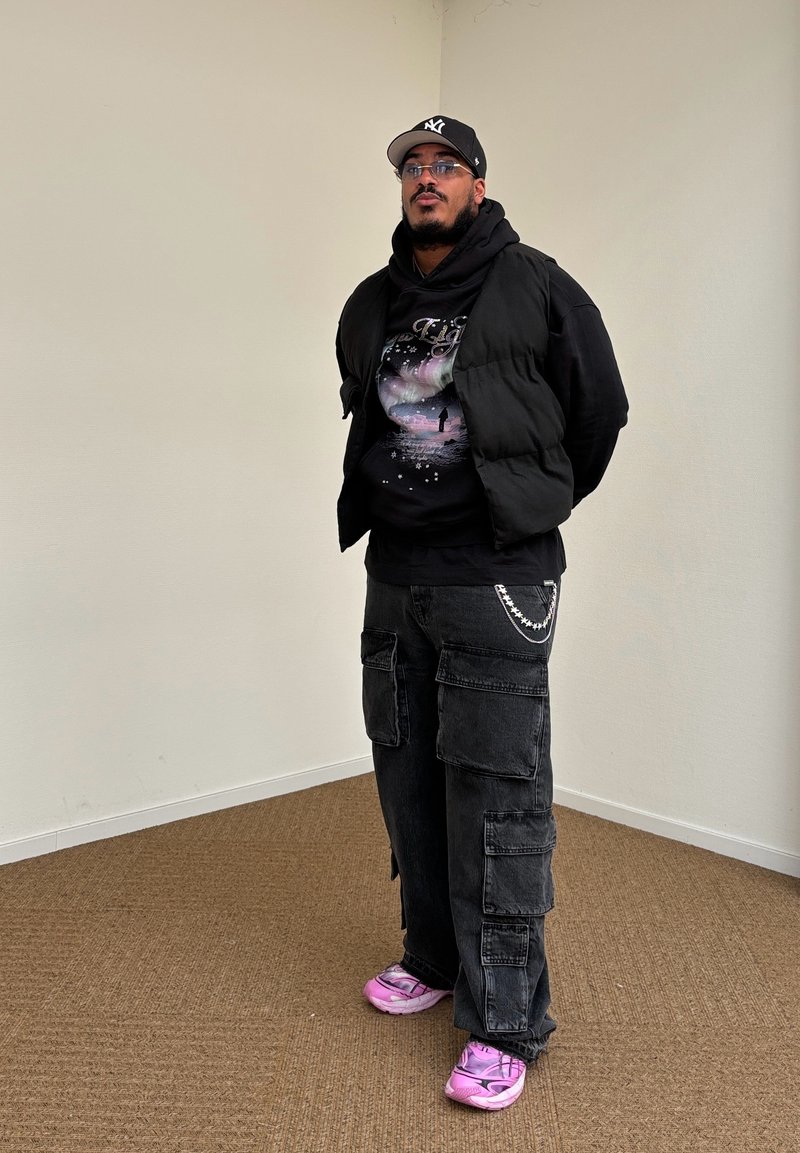 Μαύρο αμάνικο μπουφάν πάνω από μια μαύρη φούτερ με κοσμικό γραφικό. Σκοτεινά cargo παντελόνια με πολλές τσέπες και ροζ αθλητικά παπούτσια με σχεδιασμό.