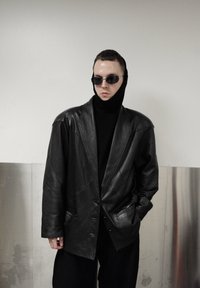 Blazer oversize en cuir noir avec épaules structurées, boutons sur le devant et poches latérales ; porté sur un col roulé noir. Lunettes de soleil noires incluses.