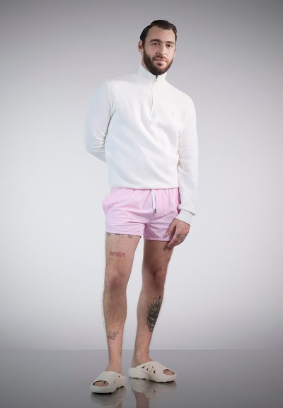 Sudadera blanca con cremallera, pantalones de baño rosas y zapatos de slip-on blancos. Tatuajes visibles en las piernas, cabello peinado hacia atrás. Fondo neutral.