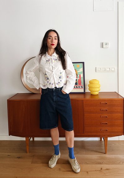 Blusa blanca con bordado floral, con cuello atado; pantalones cortos de mezclilla azul tipo bermuda; zapatos grises; calcetines azules; aparador de madera al fondo.