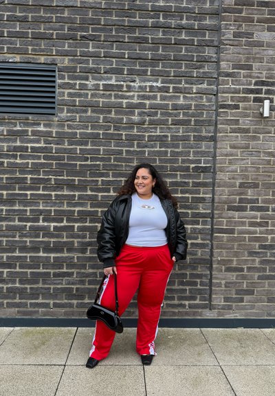 Chaqueta de plumas negra, camiseta gris, pantalones deportivos rojos con rayas blancas. Sosteniendo un pequeño bolso negro, de pie contra una pared de ladrillos.