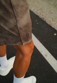 Barna futóshort, puha tapintással és logóval a szegélyen; fehér sportcipővel és zoknival viselve, sötét útfelületen.