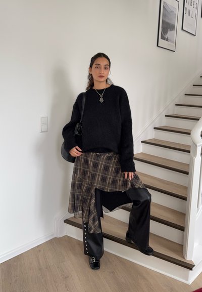 Mujer de pie en las escaleras, vistiendo un suéter negro, falda de cuadros sobre pantalones negros, zapatos negros, collar y llevando un bolso de hombro negro.