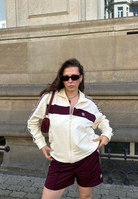 Balta rāvējslēdzēja jaka ar bordo svītru un logotipu, kopā ar bordo šortiem. Aksesuāri: maza sarkana soma un saulesbrilles.