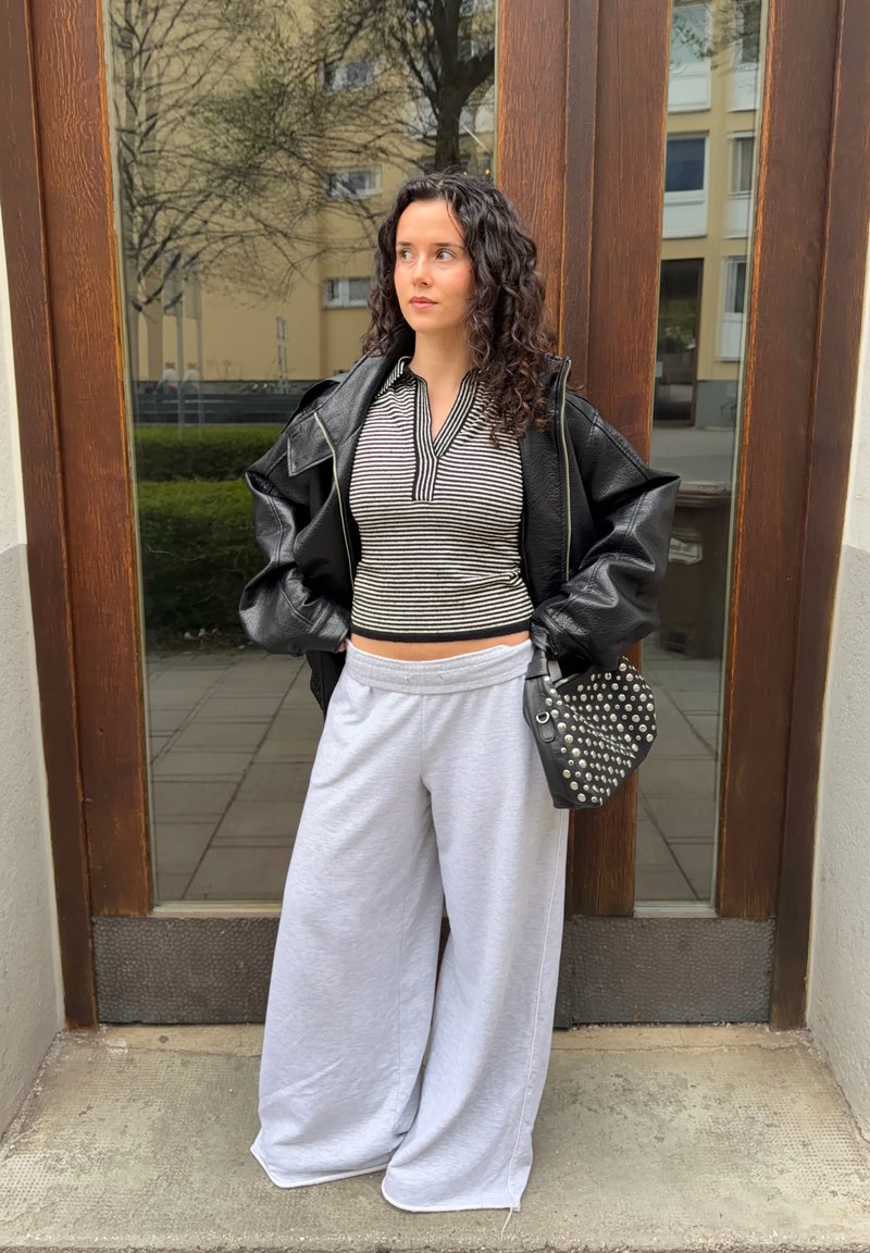 Γυναίκα με σγουρά μαλλιά, φορώντας ριγέ πουκάμισο, μαύρο δερμάτινο μπουφάν, φαρδιά λευκά παντελόνια και κρατώντας μαύρη τσάντα με τρουκς, στέκεται κοντά σε γυάλινες πόρτες.