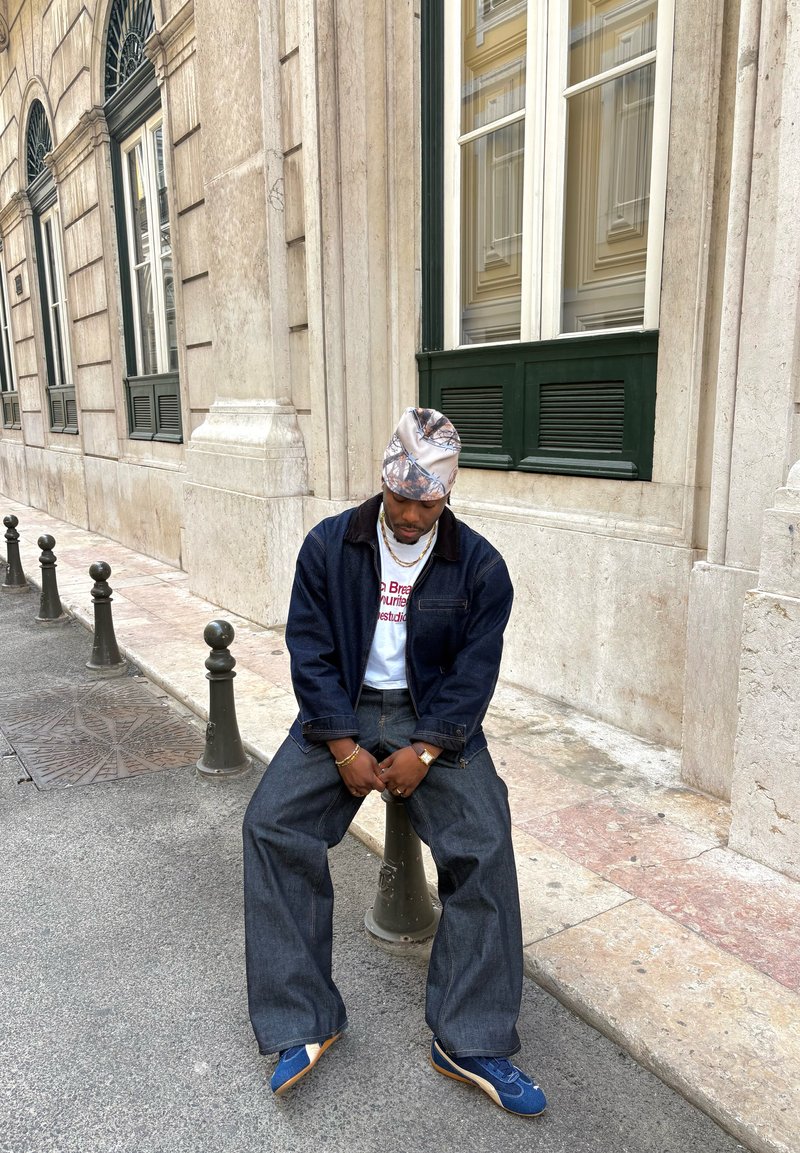 Homme portant un foulard à motifs, une veste et un pantalon en denim foncé, un t-shirt blanc avec un motif graphique, et des baskets bleues, assis sur un bollard de rue à côté d'un bâtiment en pierre.