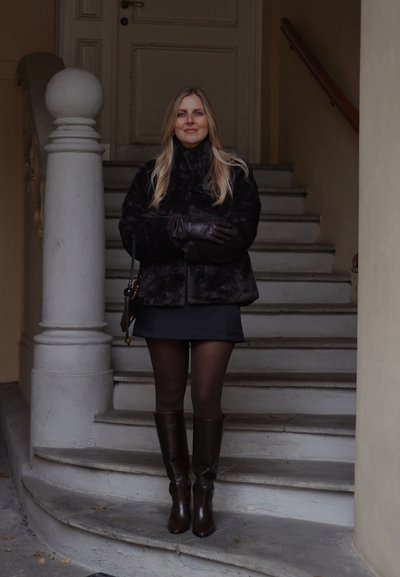 Abrigo de piel sintética negro, minifalda negra, medias negras transparentes, botas marrones hasta la rodilla. De pie en unos escalones de piedra cerca de una puerta, paredes de color marrón claro.