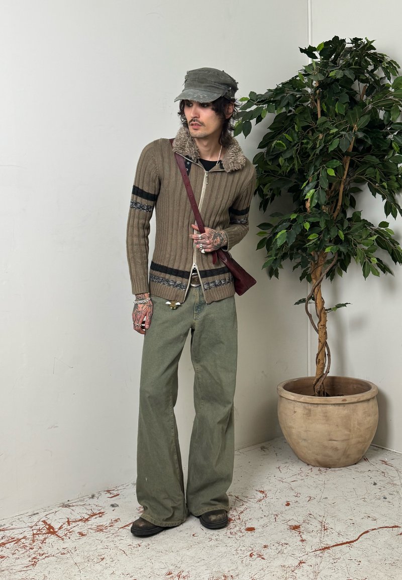 Tätoveeringutega mees, kes kannab rohelist nokamütsi, pruunide triipudega lukuga kampsunit, lahtisi rohelisi pükse ja ristkoti, seisab potitaimeste kõrval.