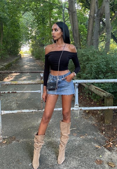 Top negro sin hombros, minifalda de mezclilla y botas beige hasta la rodilla. Pequeño bolso negro con correa de cadena. Follaje verde de fondo.