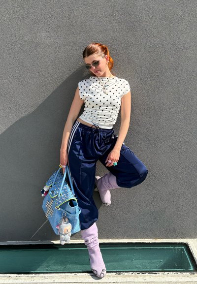 Top blanco con lunares, joggers azul marino, botas de rodilla lavanda, bolso azul con tachuelas y charms, joyería de plata, contra una pared gris.
