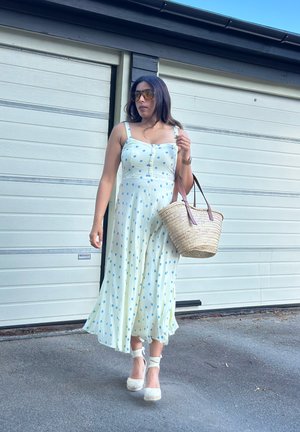 Vestido verde claro con lunares, con botones en la parte delantera, tirantes finos y falda acampanada. Espadrilles blancas y un bolso de mimbre con asas marrones.