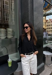 Μαύρο πουκάμισο με κουμπιά, λευκά σορτς και μαύρα γυαλιά ηλίου. Χέρια στις τσέπες. Πράσινο ποτό σε μαύρο τραπέζι στο προσκήνιο. Αστική τοποθεσία.