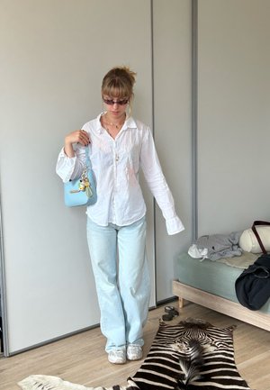 Camisa blanca de botones, jeans azul claro, zapatillas plateadas. Los accesorios incluyen un pequeño bolso azul claro con charm y gafas de sol. Alfombra de estampado de cebra.