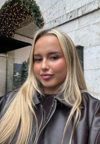 Brązowa skórzana kurtka z zamkiem z przodu, noszona na ciemnym, teksturowanym topie. Długie blond włosy i neutralny kolor ust na rozmytym tle.