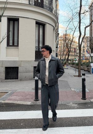 Hombre con chaqueta negra y gafas cruzando una calle de la ciudad con edificios, árboles sin hojas y pasos peatonales en el fondo.