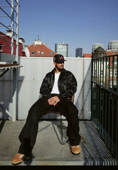 Chaqueta bomber de satén negra, camiseta blanca, pantalones anchos negros, zapatos beige. Entorno al aire libre con una barandilla de metal y fondo de ciudad.