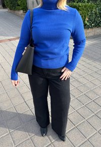 Personne portant un pull à col roulé bleu, un pantalon large noir, des chaussures noires pointues, tenant un sac à main noir sur un trottoir pavé.