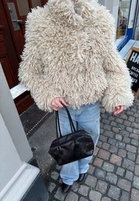 Personne portant une veste bouclée beige duveteuse, un jean bleu clair et des bottes noires, tenant un sac à main en cuir noir dans une rue pavée.