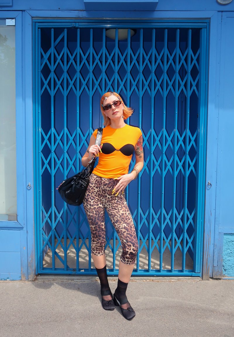 Personne portant un haut orange, un pantalon imprimé léopard, des chaussettes et des chaussures noires, debout devant une porte métallique bleue avec un sac à main noir à l'épaule.