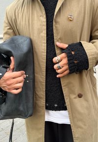 Μπεζ εξωτερικό μπουφάν με κουμπιά, μαύρος πλεκτός πουλόβε�ρ με ανοίγματα και μια μαύρη δερμάτινη τσάντα στο χέρι με μεταλλικές λεπτομέρειες και ραφές.