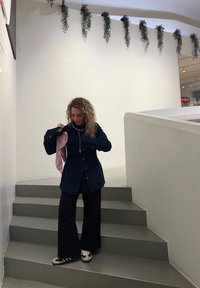 Jeansjacke kombiniert mit weiten schwarzen Hosen. Sie hält einen pinken Gegenstand und trägt eine strukturierte Halskette. Inneneinstellung mit grünen Pflanzen über der Treppe.