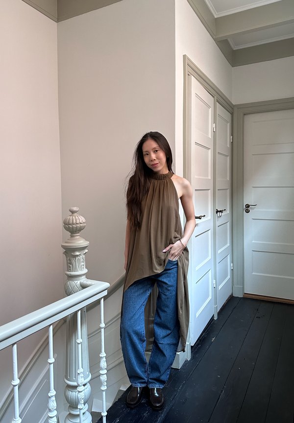 Una mujer posa en el rellano de una escalera con una camiseta sin mangas marrón, vaqueros azules y zapatos negros, junto a una barandilla blanca y unas puertas con paneles.