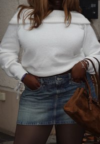 Hvit strikket genser med av skuldrene-design, kombinert med et lyseblått denimskjørt og en brun semsket skinnveske. Beinene er dekket med gjennomsiktige strømpebukser.