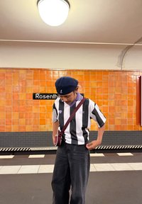Giovane vestirsi con una maglietta a righe bianco e nere e un cappellino blu navy, in piedi su una piattaforma della metropolitana con pareti piastrellate arancioni e un'insegna "Rosenthal".