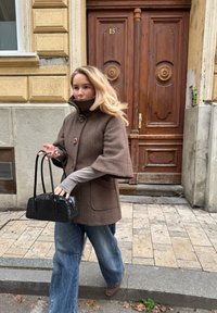 Abrigo marrón de cuadros con mangas anchas y cierre de botones, blusa gris de manga larga por debajo, jeans azules holgados, sosteniendo un pequeño bolso negro.